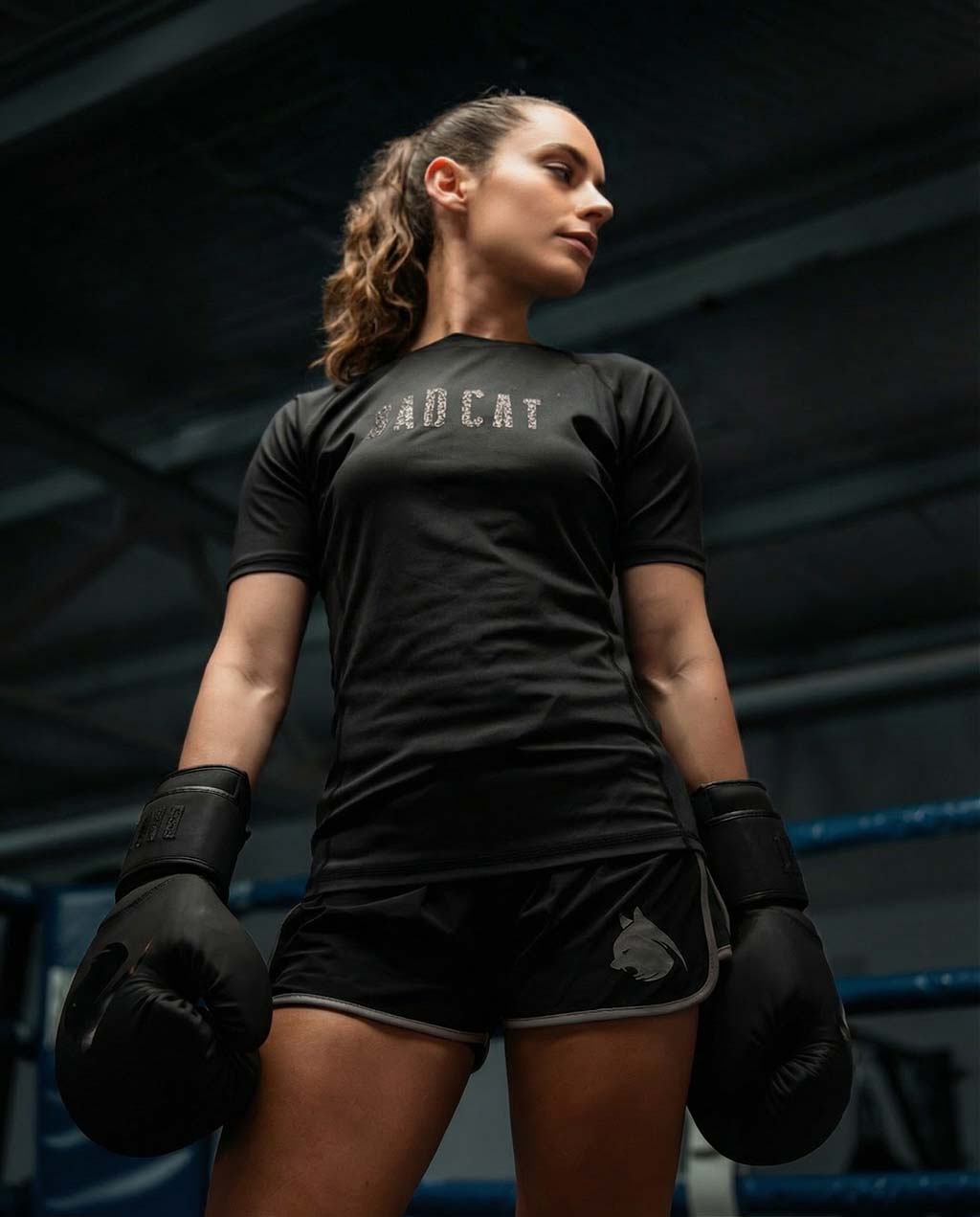 Woman in black athletic wear with 'Bad Cat' branding, holding boxing gloves in a dark setting.